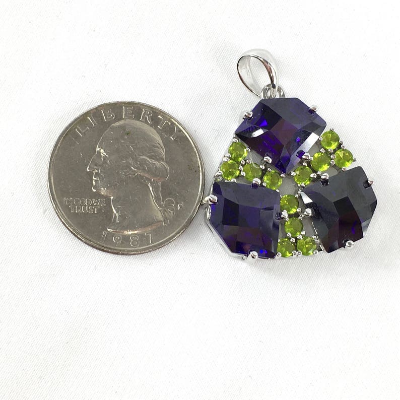 May include: A silver pendant with a triangular design featuring three large, dark purple, faceted gemstones surrounded by smaller, light green, faceted gemstones.