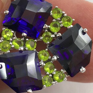 May include: A silver ring with a cluster of green and purple gemstones. The ring features a central square-shaped purple gemstone surrounded by smaller green gemstones.