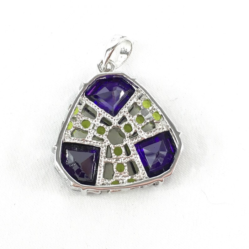 May include: A silver pendant with a triangular shape features a geometric design with four purple gemstones and smaller green gemstones.