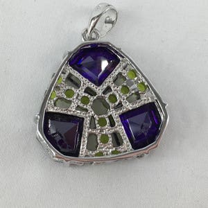May include: A silver pendant with a geometric design featuring purple and green gemstones. The pendant has a triangular shape and a bail for attaching a chain.