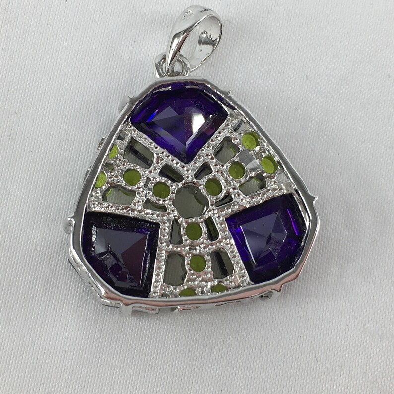 May include: A silver pendant with a geometric design featuring purple and green gemstones. The pendant has a triangular shape and a bail for attaching a chain.
