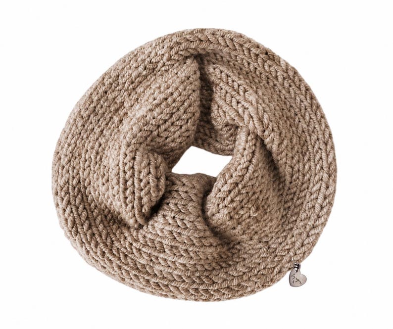 May include: A beige infinity scarf featuring a chunky knit texture with a braided pattern. The circular scarf includes a small, metallic, heart-shaped charm attached to the lower edge, adding a subtle detail to the overall knitted design.