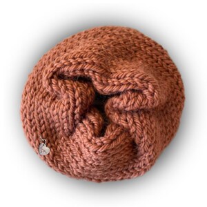 May include: A round, rust-colored knitted scrunchie made of thick yarn with a gathered, textured surface. A small, silver-toned metal heart-shaped charm is attached to the side of the knitted fabric, featuring engraved text on its surface.