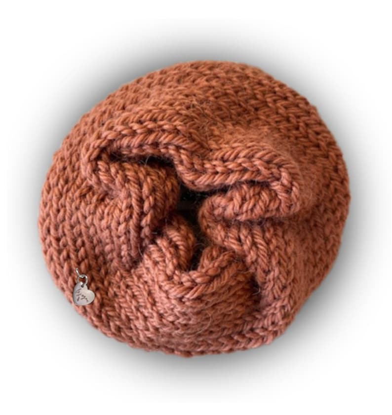 May include: A round, rust-colored knitted scrunchie made of thick yarn with a gathered, textured surface. A small, silver-toned metal heart-shaped charm is attached to the side of the knitted fabric, featuring engraved text on its surface.