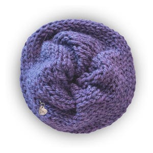 May include: A circular, hand-knitted cowl in a variegated purple yarn with a chunky, textured stitch pattern. A small, silver-toned heart-shaped metal charm is attached to the lower left side of the knitted fabric, featuring engraved script text.