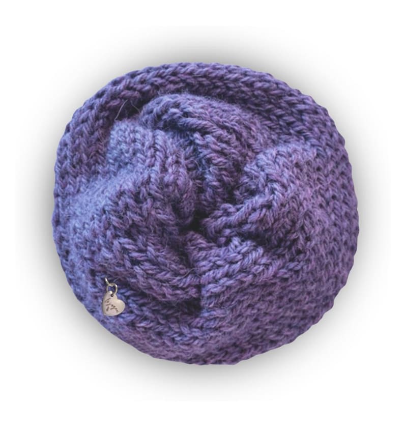 May include: A circular, hand-knitted cowl in a variegated purple yarn with a chunky, textured stitch pattern. A small, silver-toned heart-shaped metal charm is attached to the lower left side of the knitted fabric, featuring engraved script text.
