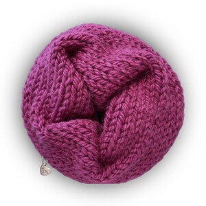 May include: A circular, magenta-colored knitted infinity scarf featuring a chunky, textured stitch pattern. A small, silver-toned heart-shaped metal charm is attached to the lower edge of the knit fabric, adding a subtle metallic detail to the design.