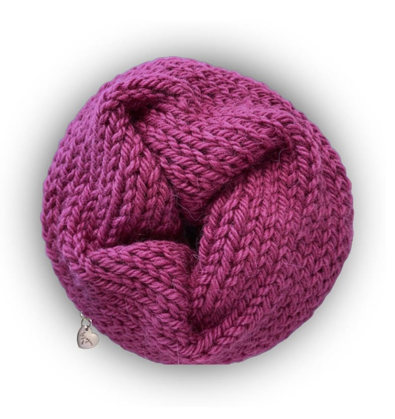 May include: A circular, magenta-colored knitted infinity scarf featuring a chunky, textured stitch pattern. A small, silver-toned heart-shaped metal charm is attached to the lower edge of the knit fabric, adding a subtle metallic detail to the design.