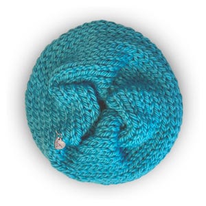 May include: A circular, hand-knitted cowl in a vibrant turquoise yarn with a thick, textured stitch pattern. A small, silver-colored heart-shaped metal charm is attached to the lower left side of the knitted fabric, featuring engraved text.
