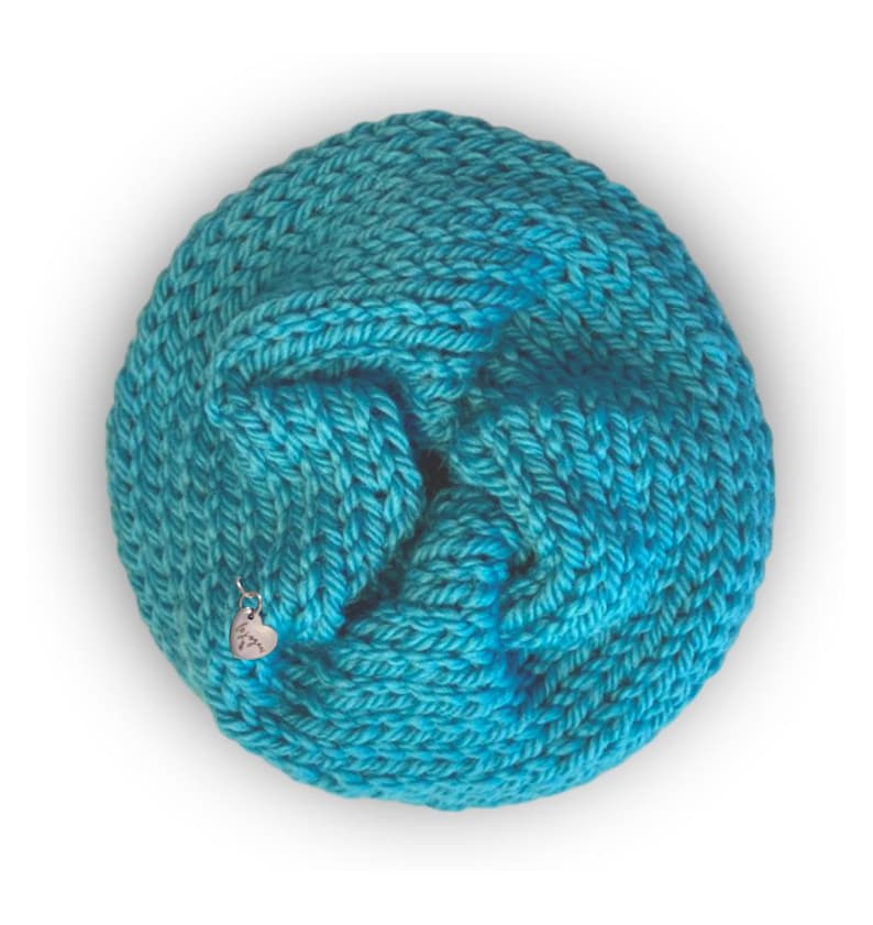 May include: A circular, hand-knitted cowl in a vibrant turquoise yarn with a thick, textured stitch pattern. A small, silver-colored heart-shaped metal charm is attached to the lower left side of the knitted fabric, featuring engraved text.