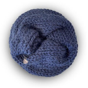 May include: A circular, dark blue knitted accessory featuring a chunky, textured yarn pattern. The item is folded into a twisted, layered shape and includes a small, silver-toned heart-shaped metal charm attached to the edge of the knit fabric.