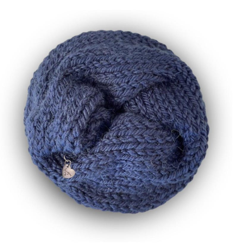 May include: A circular, dark blue knitted accessory featuring a chunky, textured yarn pattern. The item is folded into a twisted, layered shape and includes a small, silver-toned heart-shaped metal charm attached to the edge of the knit fabric.