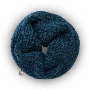 May include: A deep teal infinity scarf featuring a chunky knit texture with a braided pattern. The circular scarf is adorned with a small, silver-toned metallic heart-shaped charm attached to the lower edge, adding a subtle detail to the design.