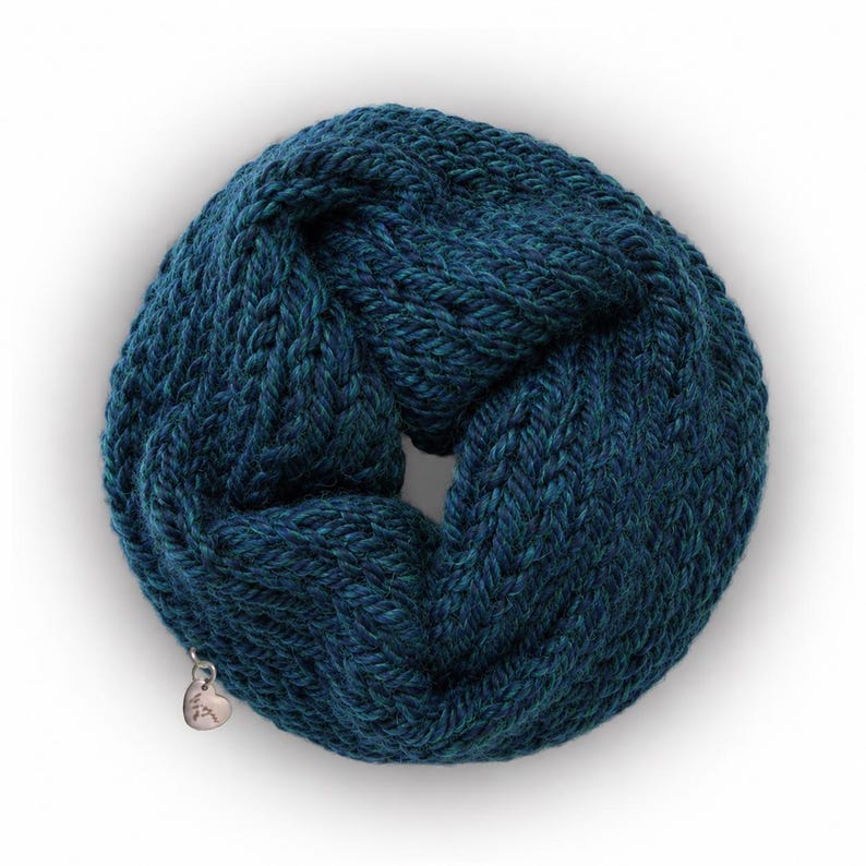 May include: A deep teal infinity scarf featuring a chunky knit texture with a braided pattern. The circular scarf is adorned with a small, silver-toned metallic heart-shaped charm attached to the lower edge, adding a subtle detail to the design.