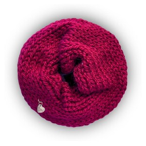 May include: A circular, magenta-colored knitted infinity scarf with a thick, textured yarn construction. A small, silver-toned metal heart-shaped charm is attached to the lower left side of the knitted fabric, featuring engraved text.