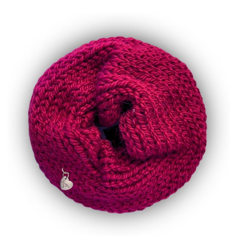 May include: A circular, magenta-colored knitted infinity scarf with a thick, textured yarn construction. A small, silver-toned metal heart-shaped charm is attached to the lower left side of the knitted fabric, featuring engraved text.