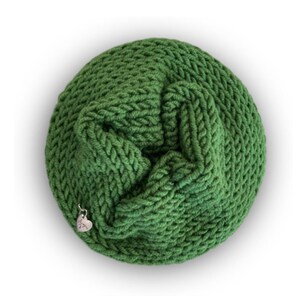 May include: A circular, forest green knitted cowl with a thick, textured yarn weave. A small, silver-toned heart-shaped metal charm is attached to the lower edge of the knit fabric, featuring a subtle engraved design on its surface.