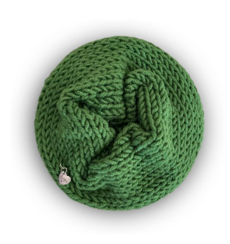 May include: A circular, forest green knitted cowl with a thick, textured yarn weave. A small, silver-toned heart-shaped metal charm is attached to the lower edge of the knit fabric, featuring a subtle engraved design on its surface.
