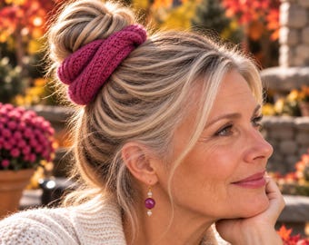 May include: A magenta knitted hair scrunchie wrapped around a high bun and a matching drop earring featuring a central magenta bead flanked by two small white pearls on a gold-toned metal hook, worn by a person with blonde hair.