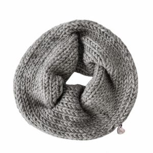 May include: A chunky knit infinity scarf in a solid light gray color. The scarf features a thick, textured yarn construction with a circular loop design. A small, metallic heart-shaped charm is attached to the lower edge of the knitted fabric.