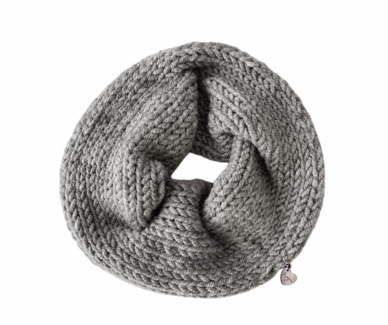 May include: A chunky knit infinity scarf in a solid light gray color. The scarf features a thick, textured yarn construction with a circular loop design. A small, metallic heart-shaped charm is attached to the lower edge of the knitted fabric.