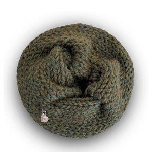 May include: A circular, hand-knitted cowl in a variegated olive green yarn with a chunky, textured stitch pattern. A small, silver-toned metal heart-shaped charm is attached to the lower left side of the knitted fabric, adding a subtle detail.
