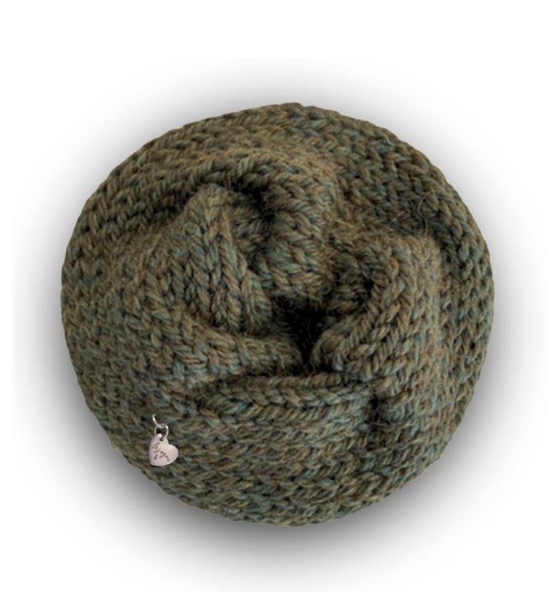 May include: A circular, hand-knitted cowl in a variegated olive green yarn with a chunky, textured stitch pattern. A small, silver-toned metal heart-shaped charm is attached to the lower left side of the knitted fabric, adding a subtle detail.