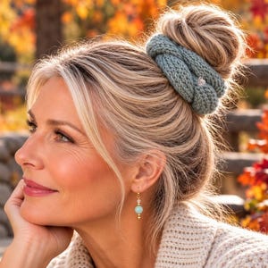 May include: A sage green knitted hair scrunchie wrapped around a bun and a single gold-toned drop earring featuring a small clear bead above a round, light blue-green stone bead, suspended from a gold hook wire.