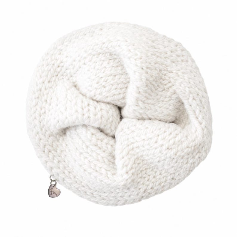 May include: A round, off-white knitted accessory with a thick, textured yarn construction. The item features a small, metallic, heart-shaped charm attached to the edge by a silver-toned jump ring, adding a subtle detail to the circular knit piece.