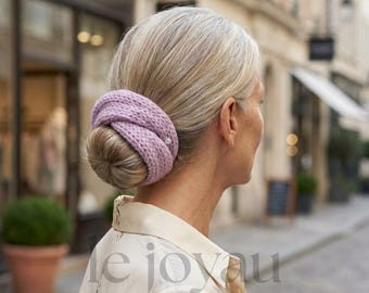 May include: A woman with light gray hair styled in a bun, adorned with a lavender knitted hair accessory. The accessory is wrapped around the bun, adding a touch of color and texture. The background shows a blurred street scene.