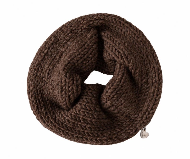 May include: A dark brown infinity scarf featuring a chunky knit texture with a circular loop design. A small, silver-toned heart-shaped metal charm is attached to the lower edge of the scarf, adding a subtle metallic detail to the dark brown yarn.