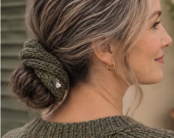 May include: An olive green knitted hair scrunchie wrapped around a hair bun. The accessory features a textured, chunky knit pattern and a small, silver-toned heart-shaped charm dangling from the side. The knit fabric is a solid, deep olive green.