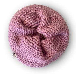 May include: A circular, dusty rose-colored knitted accessory with a thick, textured yarn construction. The piece features a folded, layered design and includes a small, silver-toned heart-shaped metal charm attached to the outer edge.