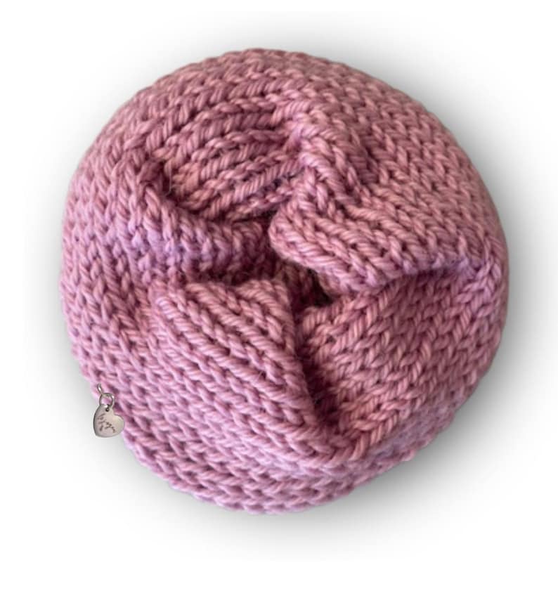 May include: A circular, dusty rose-colored knitted accessory with a thick, textured yarn construction. The piece features a folded, layered design and includes a small, silver-toned heart-shaped metal charm attached to the outer edge.