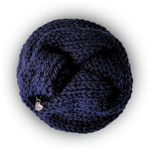 May include: A circular, dark navy blue knitted accessory featuring a thick, textured yarn construction with a braided or twisted pattern. A small, silver-toned heart-shaped metal charm is attached to the edge of the knit fabric.