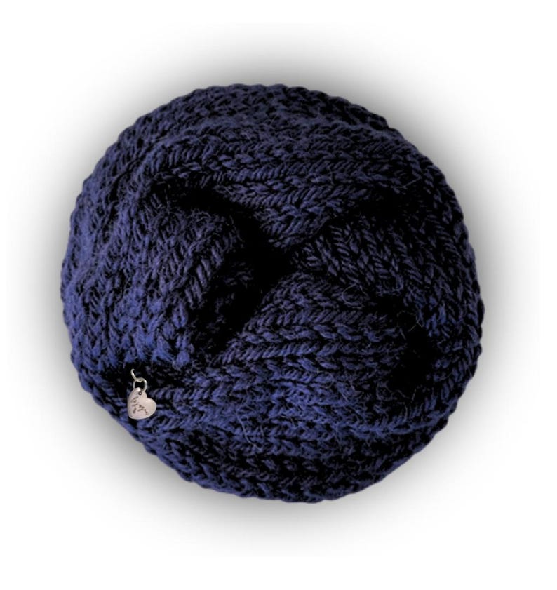 May include: A circular, dark navy blue knitted accessory featuring a thick, textured yarn construction with a braided or twisted pattern. A small, silver-toned heart-shaped metal charm is attached to the edge of the knit fabric.