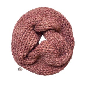 May include: A chunky knit infinity scarf in a dusty rose color featuring a thick, braided cable pattern throughout the fabric. A small, silver-toned metal charm in the shape of a heart is attached to the lower edge of the circular scarf.