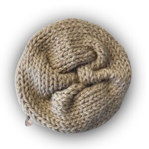 May include: A round, beige, hand-knitted pouch featuring a thick, textured yarn construction with a visible knot-like pattern on the front surface. A small, metallic, heart-shaped zipper pull is attached to the side of the circular knitted bag.