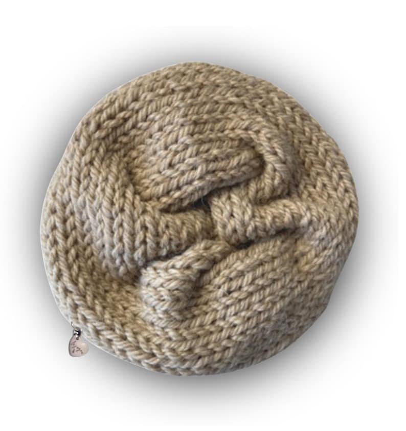 May include: A round, beige, hand-knitted pouch featuring a thick, textured yarn construction with a visible knot-like pattern on the front surface. A small, metallic, heart-shaped zipper pull is attached to the side of the circular knitted bag.