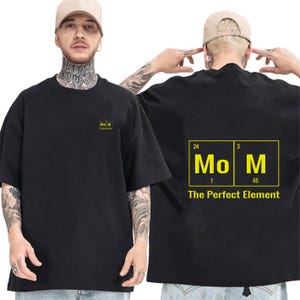 May include: A black short-sleeve t-shirt featuring a small yellow graphic on the front chest and a large yellow periodic table-style graphic on the back. The back design displays the text Mo M with the phrase The Perfect Element underneath.
