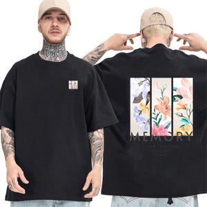 May include: A black short-sleeved t-shirt featuring a small floral graphic on the front left chest. The back displays three vertical rectangular panels containing colorful floral illustrations with the text MEMORY NEVER FADE AWAY THROUGH TIMES.