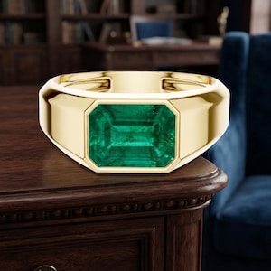 Può includere: Un anello in oro con una grande pietra preziosa verde a taglio smeraldo rettangolare incastonata al centro. La fascia è in oro giallo liscio e lucido con un profilo ampio e piatto che si restringe leggermente verso il retro, esaltando la pietra.