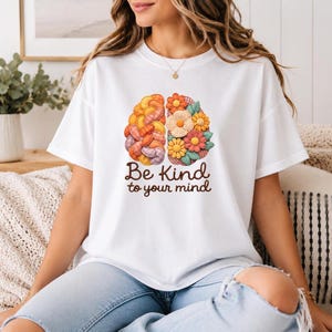 Può includere: T-shirt bianco con grafica a forma di cervello colorato con elementi floreali e la scritta "Be Kind to your mind" in marrone. Il disegno è centrato sulla parte anteriore della maglietta.