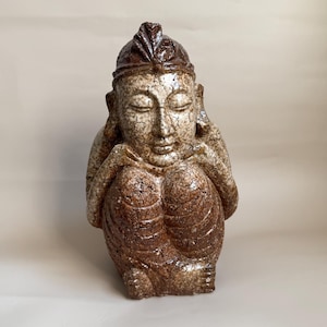 Könnte beinhalten: Eine Keramikskulptur einer sitzenden Figur mit geschlossenen Augen, die das Kinn auf die Hände stützt. Die Figur hat eine glänzende, rissige Glasur in Beige-, Braun- und Beigetönen sowie einen dunkelbraunen, fächerförmigen Kopfschmuck.