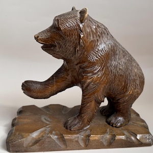 Könnte beinhalten: Eine dunkelbraune Holzfigur eines Bären, der auf einem rechteckigen Sockel steht. Der Bär ist im Seitenprofil mit erhobener rechter Tatze dargestellt und weist eine detaillierte Fellstruktur sowie geschnitzte Merkmale auf.