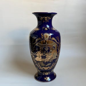 Może przedstawiać: Wazon z głębokiego kobaltowego porcelany z misternymi złotymi wzorami filigranowymi i centralnym kartuszem przedstawiającym szczegółową scenę postaci we wnętrzu. Wazon ma rozszerzany brzeg, smukłą szyjkę i podstawę w formie cokołu.