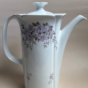 Könnte beinhalten: Eine weiße Porzellan-Kaffeekanne mit Deckel, Henkel und Ausguss. Die Kanne ist mit einem floralen Muster in Lila- und Grüntönen verziert. Die Kanne hat ein Fassungsvermögen von ca. 950 ml.