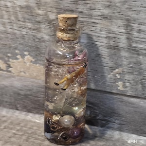 May include: A clear glass bottle with a cork stopper filled with a transparent liquid, containing a small miniature figure in a yellow outfit, several white spherical beads, and various metallic charms suspended within the fluid.