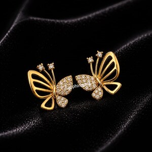 Peut inclure: Une paire de boucles d'oreilles papillon dorées. Chaque boucle d'oreille présente un motif de papillon, avec une aile recouverte de petites pierres scintillantes. Les antennes de chaque papillon sont ornées de trois pierres supplémentaires.