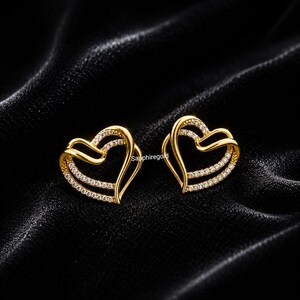 Peut inclure: Une paire de boucles d'oreilles dorées en forme de cœur. Chaque boucle d'oreille présente un double cœur orné d'une rangée de petits diamants transparents. Les boucles d'oreilles sont posées sur un fond noir texturé.